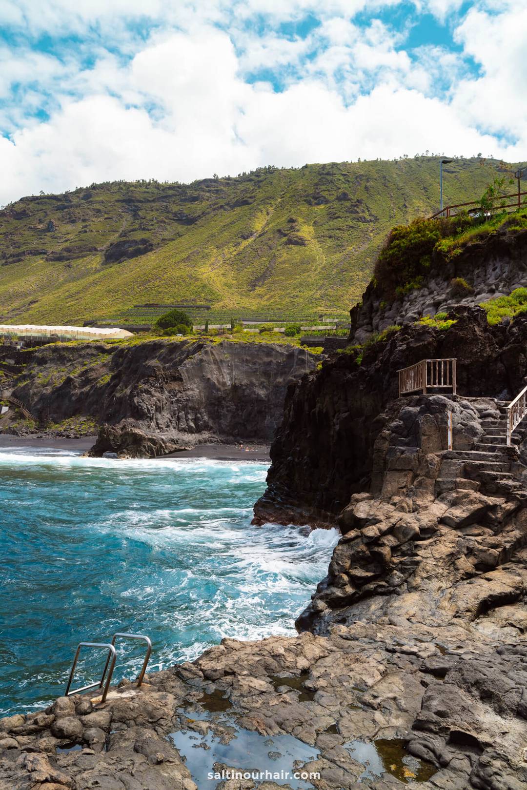 Rocky coastline with metal stairs leading into the ocean on La Palma, waves crashing against the rocks, and green hills in the background under a partly cloudy sky&mdash;an iconic scene from any Canary Islands travel guide.