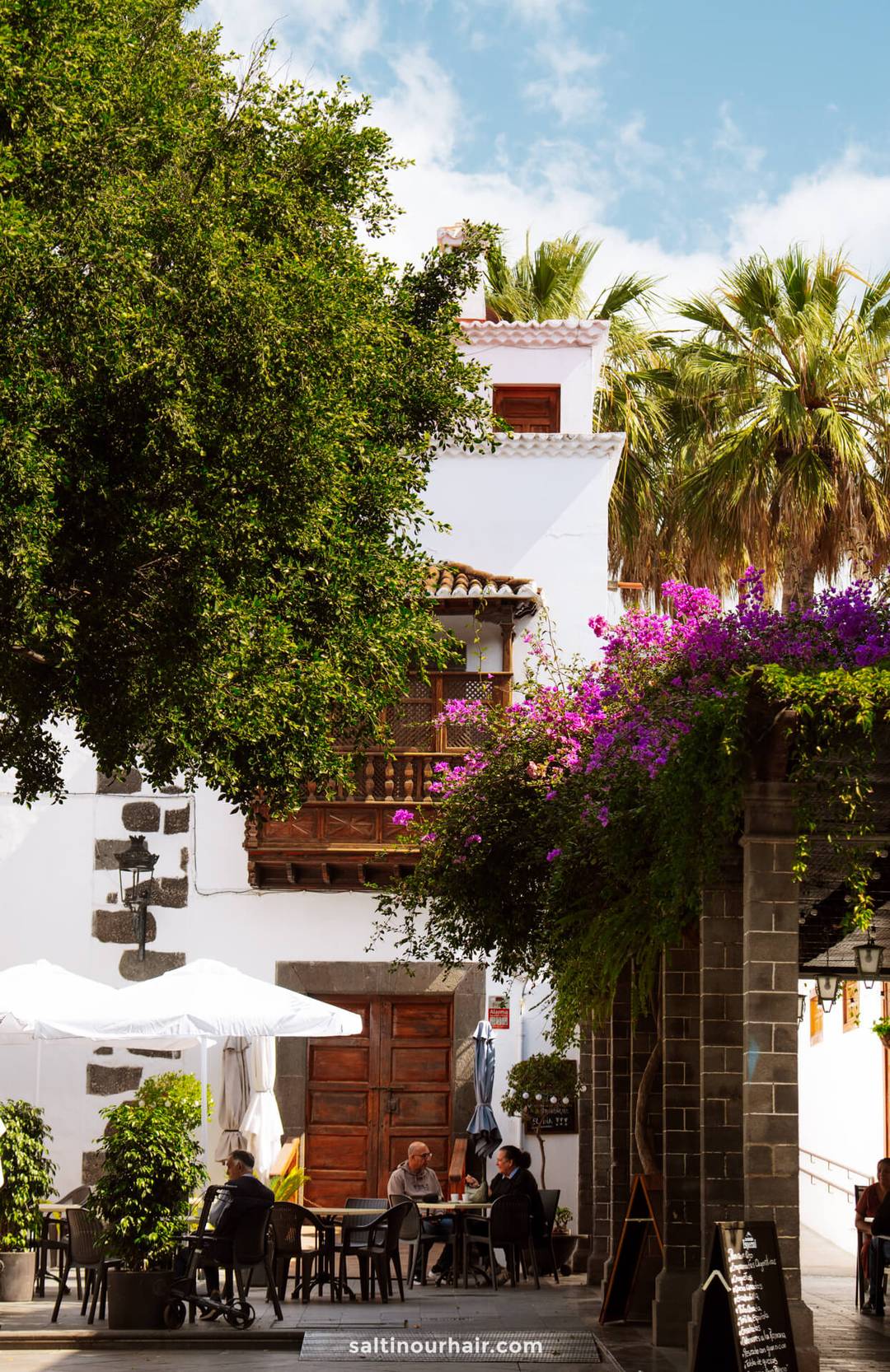 People sit at tables under umbrellas in a shaded outdoor caf&eacute; on La Palma, surrounded by greenery and blooming flowers, with a white building and palm trees in the background&mdash;a perfect scene from any Canary Islands travel guide.