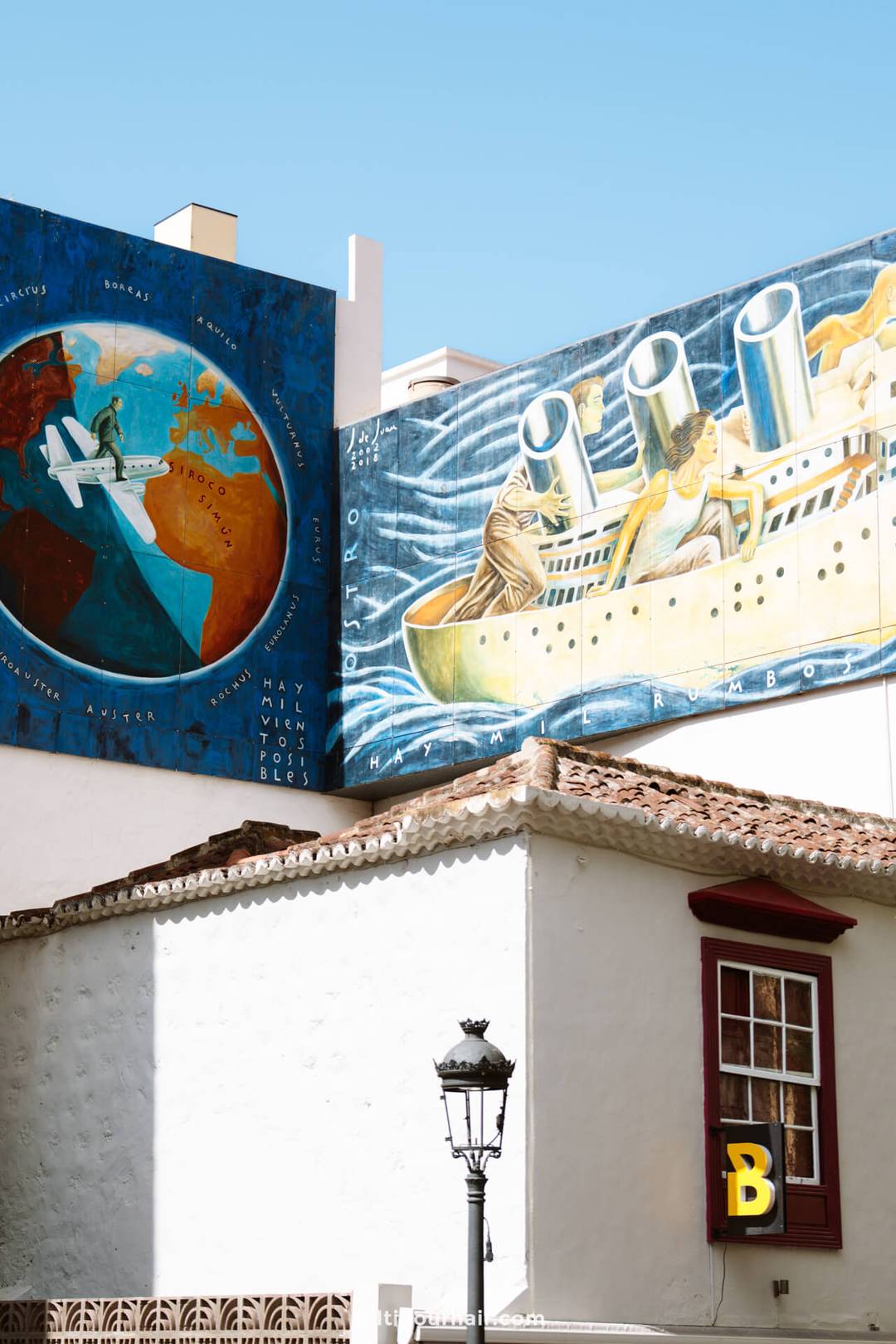 Two colorful murals on a building wall in La Palma: one shows an airplane over a globe, the other depicts a large ship on water. A white building with a red window and a yellow B is in the foreground&mdash;perfect for any Canary Islands travel guide.