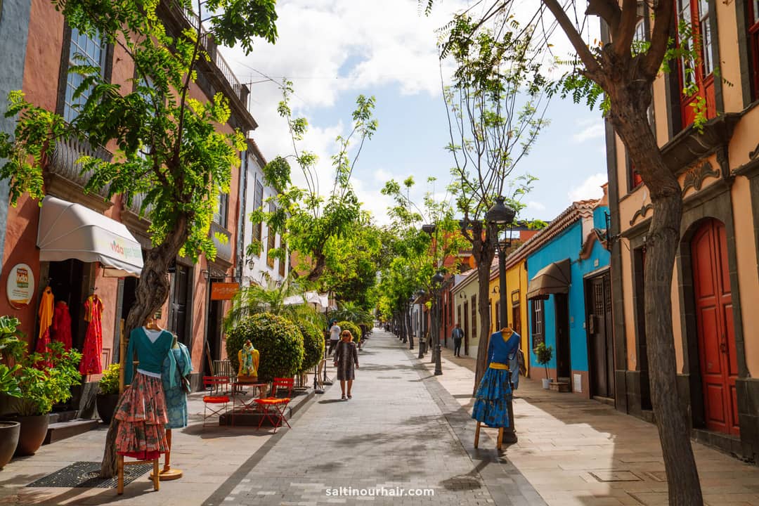 Pedestrian street in La Palma, Canary Islands, lined with colorful buildings, outdoor seating, mannequins in dresses, and trees; a few people stroll the pathway under a partly cloudy sky&mdash;a scene fit for any travel guide.