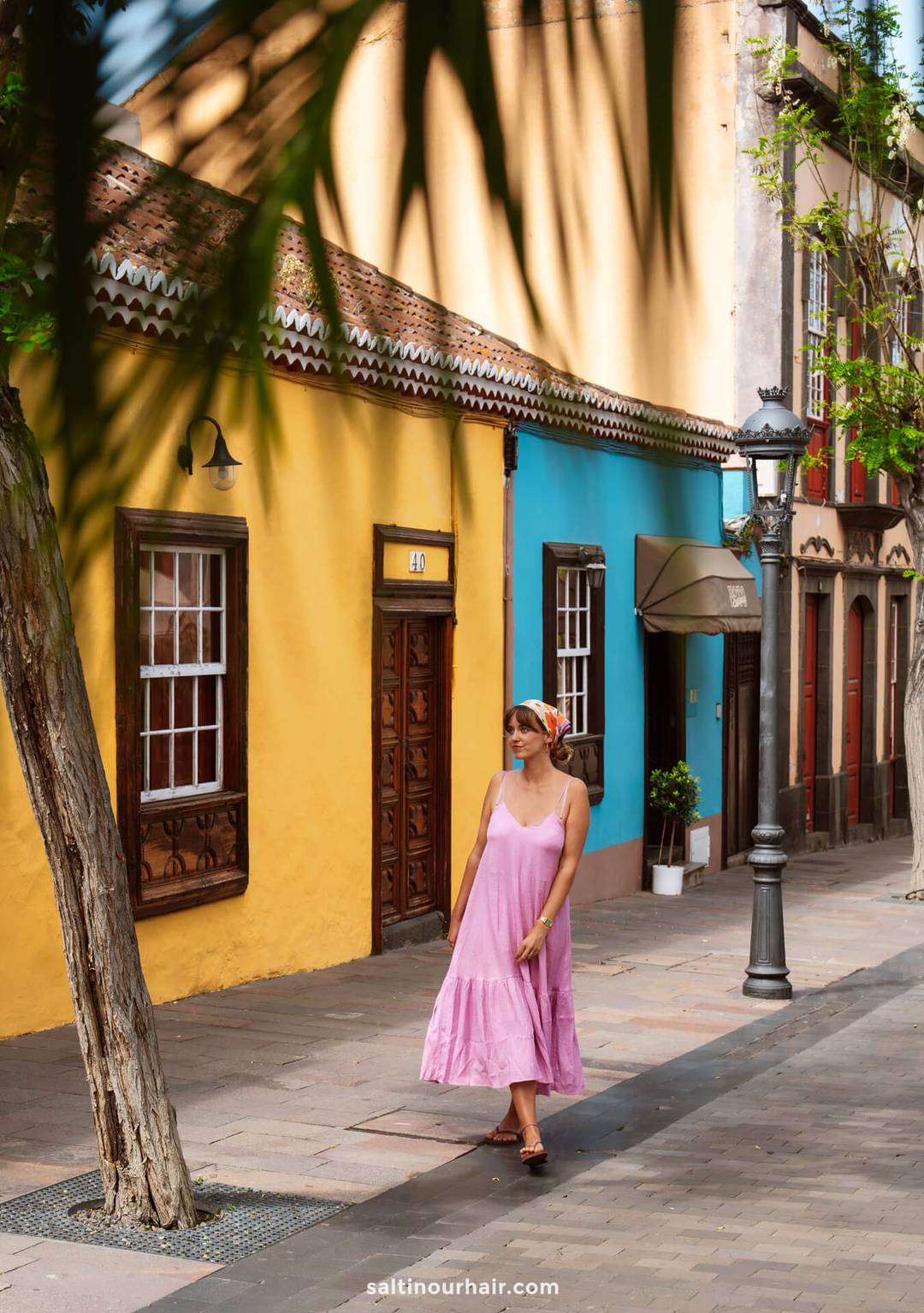 A woman in a pink dress and headscarf strolls along a sunlit street in La Palma, with colorful yellow and blue buildings, trees, and street lamps&mdash;perfect for any Canary Islands travel guide.
