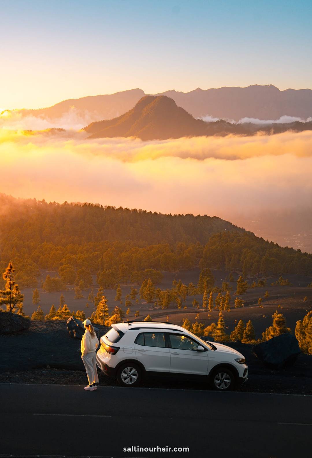 A person stands next to a white SUV parked on a mountain road at sunrise in La Palma, overlooking a valley filled with clouds and surrounded by forested hills in the Canary Islands.