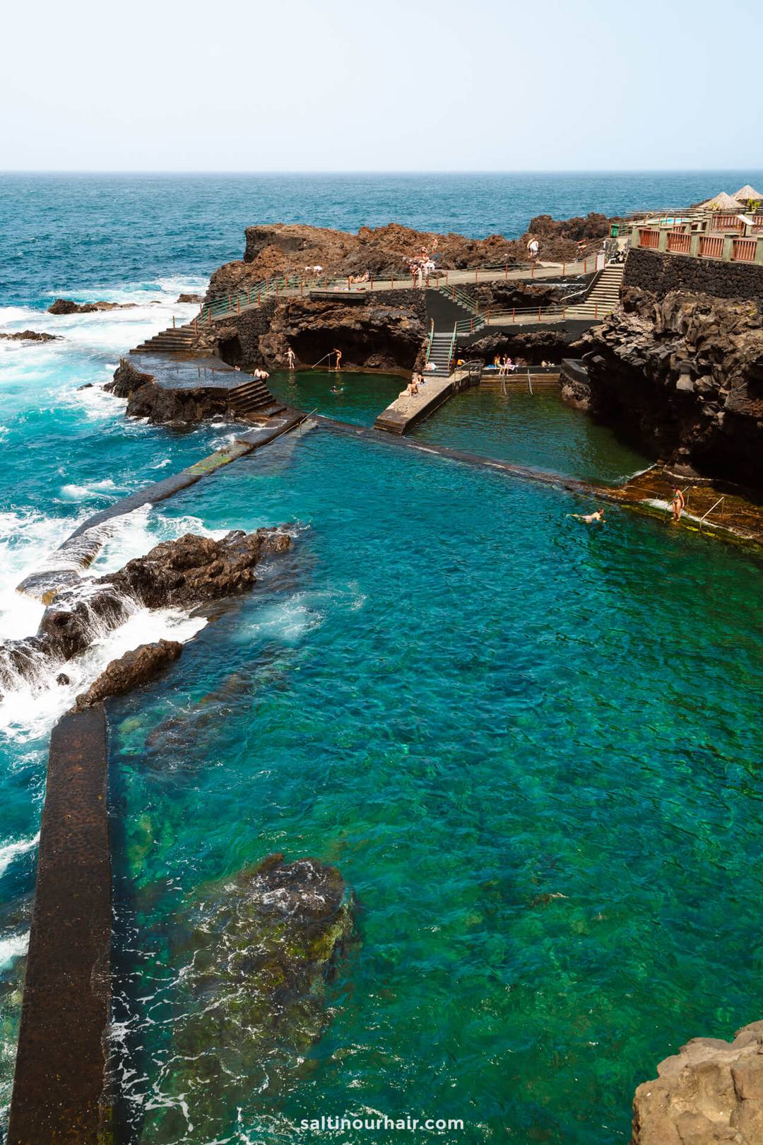 Natural volcanic rock pools with clear turquoise water, surrounded by rocky cliffs and people swimming, near the ocean under a clear sky&mdash;La Palma&rsquo;s hidden gem in the Canary Islands, perfect for your next travel guide adventure.