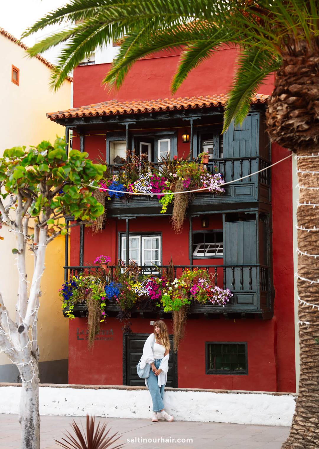 A woman stands in front of a red building in La Palma, its two wooden balconies adorned with colorful flowers and plants&mdash;a charming scene from this Canary Islands travel guide.