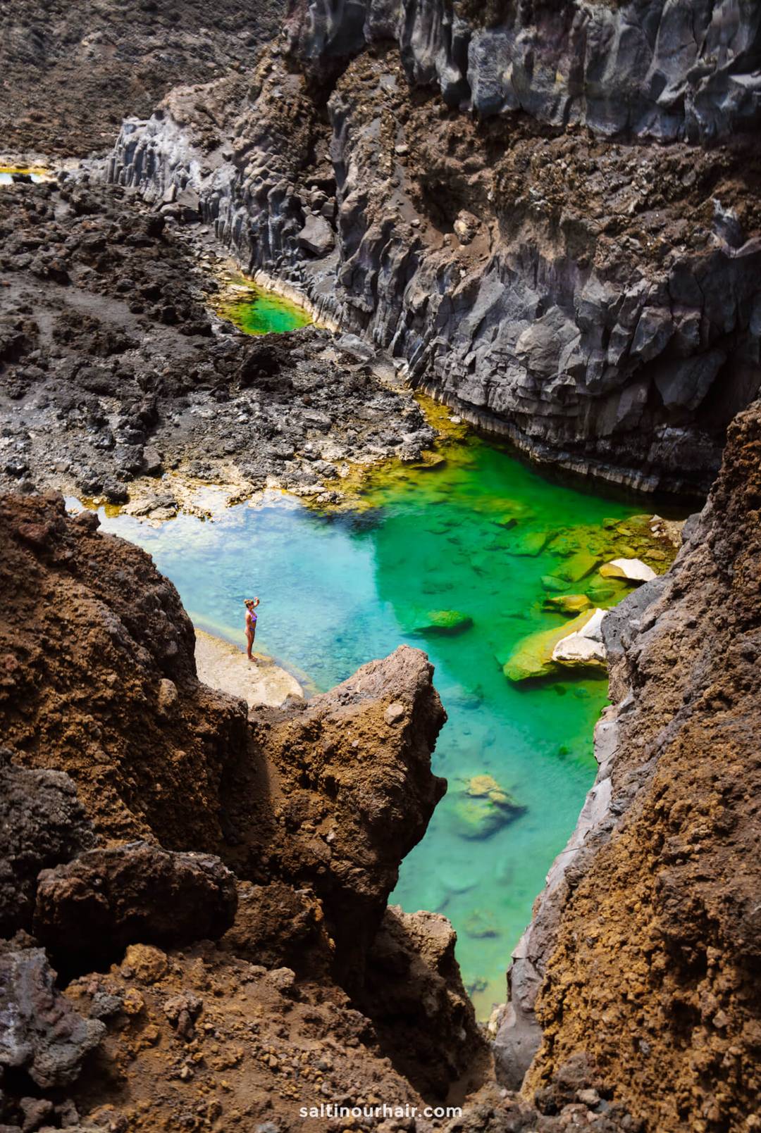 A person stands on rocky terrain beside a clear, turquoise stream surrounded by jagged, dark cliffs&mdash;a hidden gem in La Palma, one of the Canary Islands.