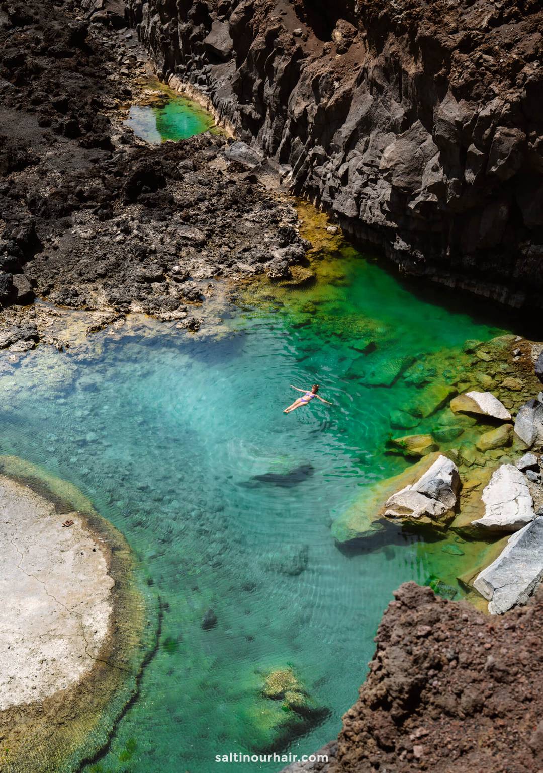 A person floats in a clear turquoise pool on La Palma, surrounded by rocky cliffs and dark volcanic terrain&mdash;a hidden gem of the Canary Islands featured in any top travel guide.