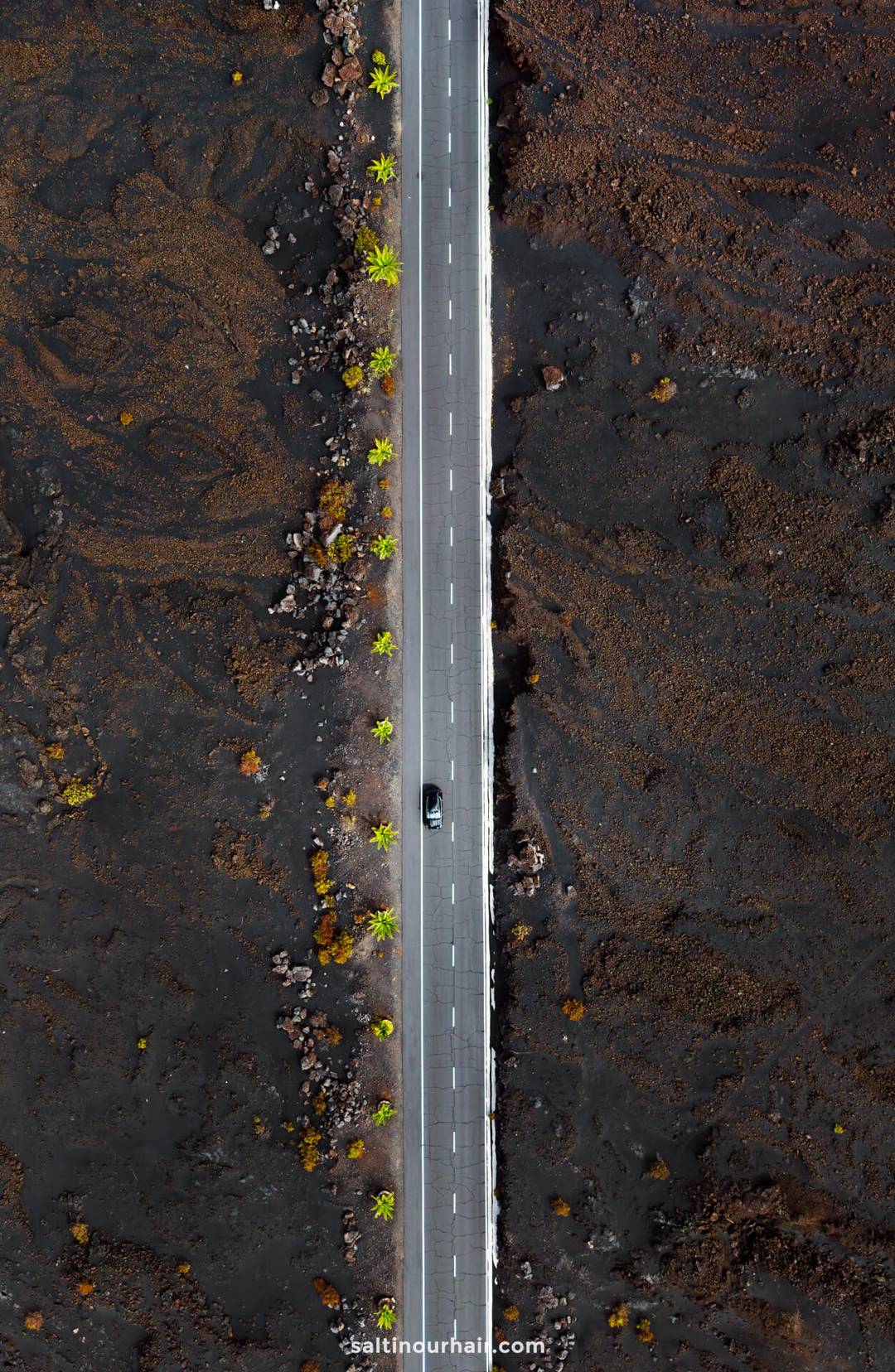 Aerial view of a straight road cutting through a barren, dark landscape on La Palma, Canary Islands, with small green trees lining both sides and a single car driving along it&mdash;perfect inspiration for your next travel guide.