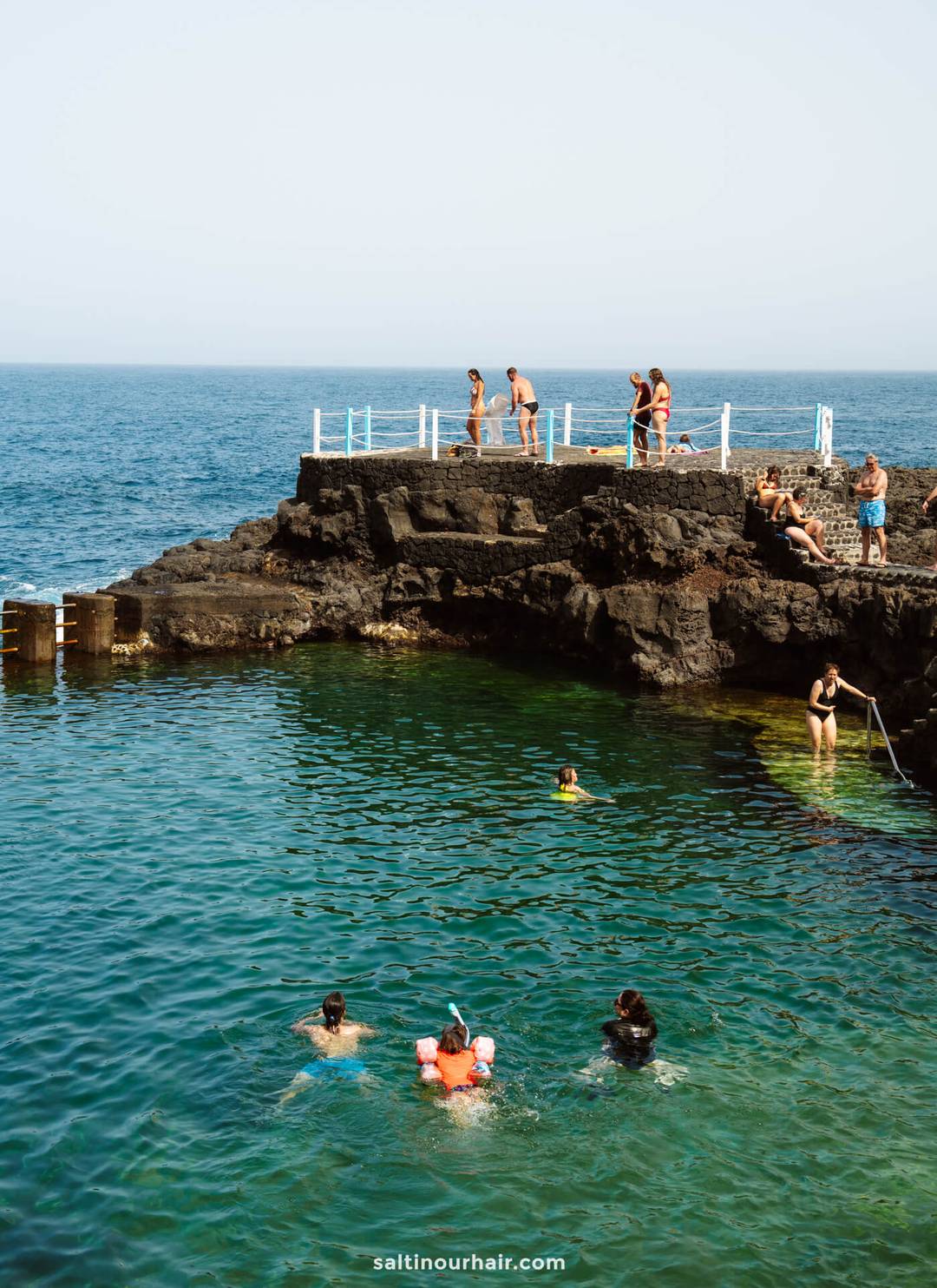 People swim and relax in a natural rock pool by the ocean on La Palma, with some climbing and jumping from the rocks; calm, clear water and a blue sky create a perfect scene from a Canary Islands travel guide.