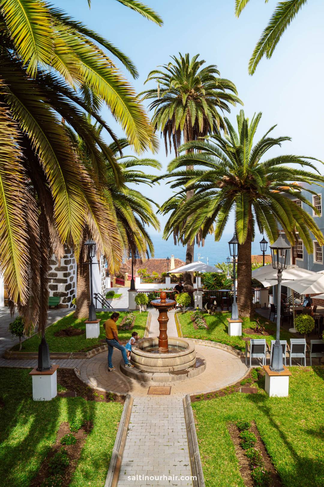 A man walks with a child near a circular fountain surrounded by palm trees and colonial-style buildings on a sunny day in La Palma, Canary Islands&mdash;a perfect snapshot for any travel guide.