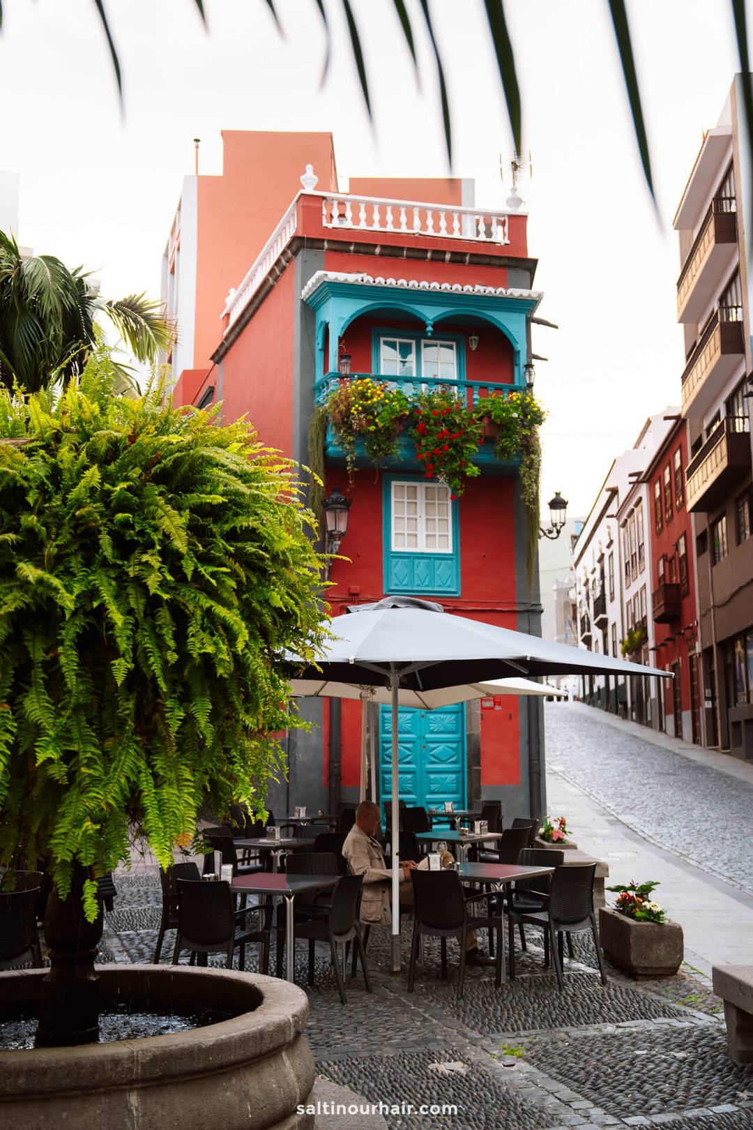 A red building with blue balconies and doors sits at a street corner; cafe tables with an umbrella and plants are in the foreground, echoing the vibrant charm of La Palma in the Canary Islands&mdash;perfect for your next travel guide inspiration.