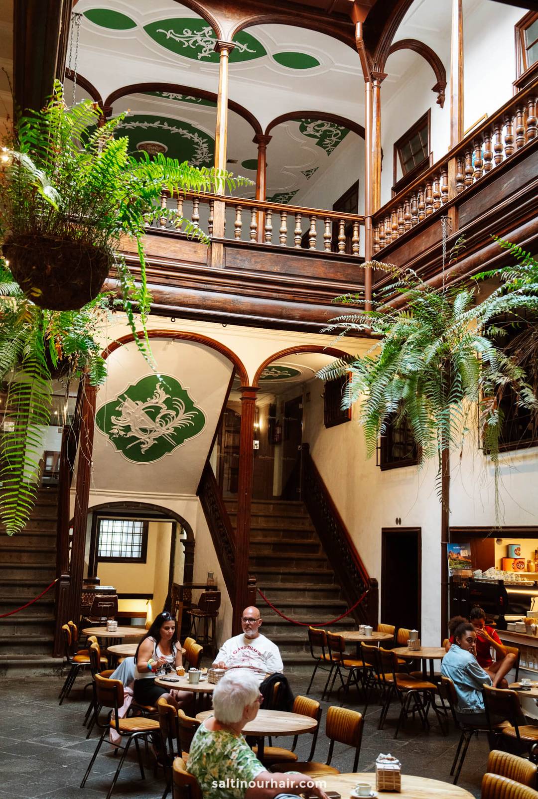 Spacious indoor courtyard with wooden balconies, green ceiling art, hanging ferns, and people seated at tables in a caf&eacute; setting&mdash;ideal for a La Palma travel guide to the hidden gems of the Canary Islands.