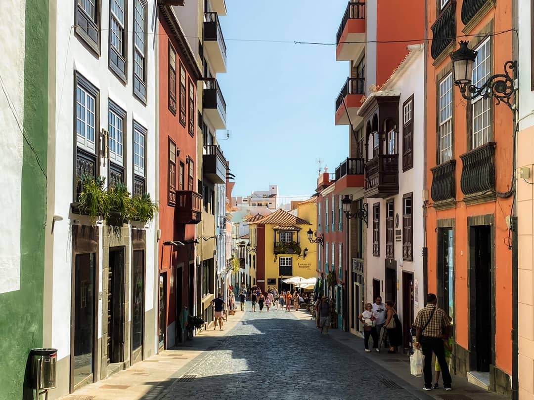 A narrow cobblestone street lined with colorful buildings in La Palma, Canary Islands, welcomes pedestrians strolling along the sidewalks, leading to a bright yellow building at the street&rsquo;s end under a clear sky&mdash;a scene fit for any travel guide.
