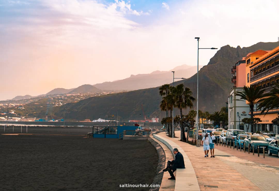 A man sits on a bench by a paved walkway next to a black sand beach in La Palma, Canary Islands, with mountains and buildings in the background under a hazy sky&mdash;a perfect scene from any travel guide.