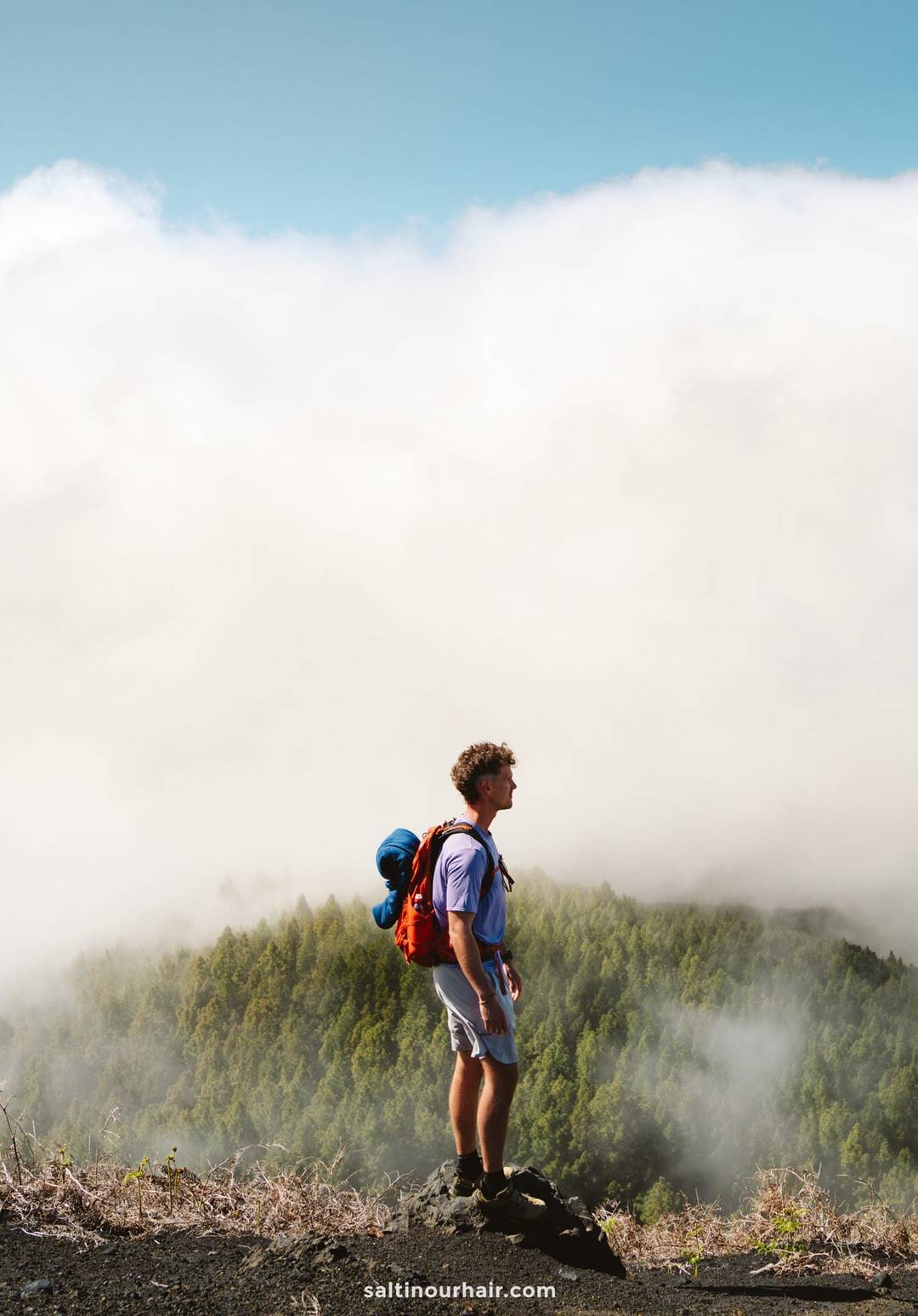 A man with a red backpack stands on a rocky ledge overlooking La Palma&rsquo;s forested landscape, clouds drifting beneath a bright blue sky&mdash;a perfect scene for any Canary Islands travel guide.