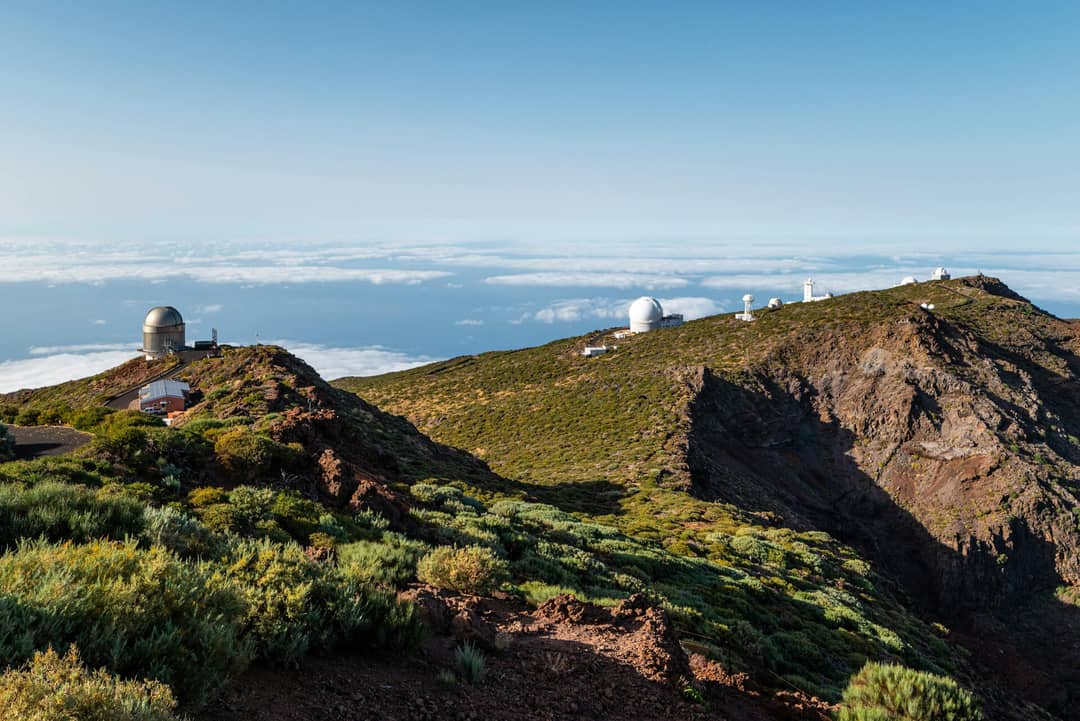 Several observatory domes sit atop a green, rocky mountain ridge under a clear sky on La Palma, one of the Canary Islands, with clouds visible in the distance&mdash;a perfect scene for any travel guide.