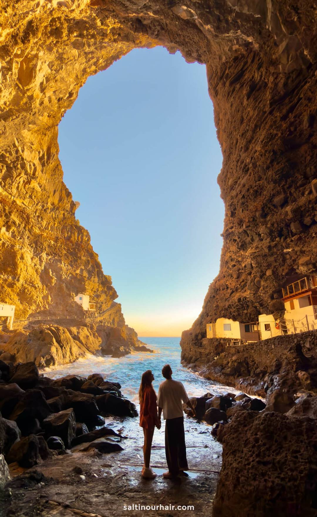 Two people stand inside a large rocky cave on La Palma, holding hands and looking out towards the sea, with sunlight illuminating the cave walls and small buildings nearby&mdash;a perfect moment in the Canary Islands for any travel guide.