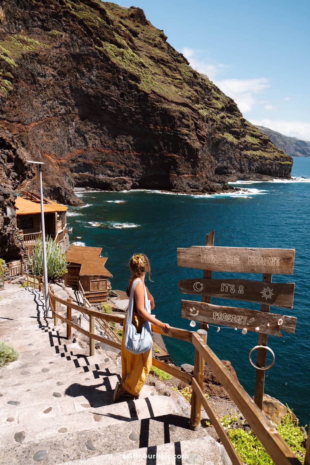 A woman stands on stone steps overlooking the ocean on La Palma in the Canary Islands, near a wooden signpost and rustic houses built into a steep, rocky cliff&mdash;a perfect scene for any travel guide.