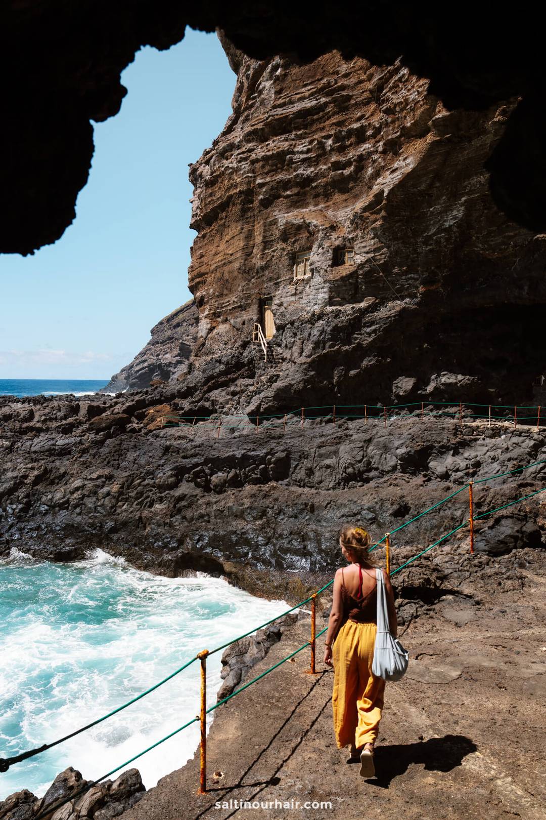 A person walks along a rocky coastal path in La Palma, Canary Islands, with waves crashing below and cliffs rising under a partly cloudy sky&mdash;a perfect scene from a travel guide.