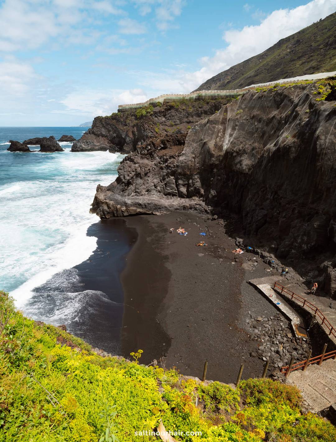 Tucked away on La Palma in the Canary Islands, this small black sand beach is surrounded by high rocky cliffs and waves, with a few people sunbathing and a wooden walkway leading down to the shore&mdash;a hidden gem for any travel guide.