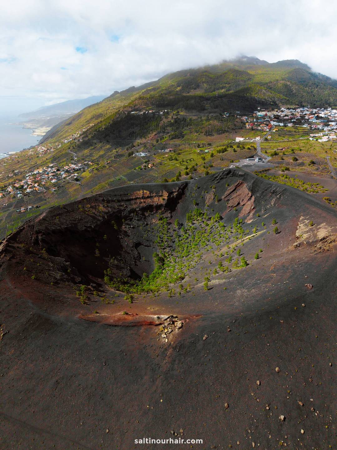 Aerial view of a large volcanic crater with sparse vegetation on La Palma, Canary Islands, surrounded by hills, houses, and a distant coastline under a partly cloudy sky&mdash;perfect for any travel guide.