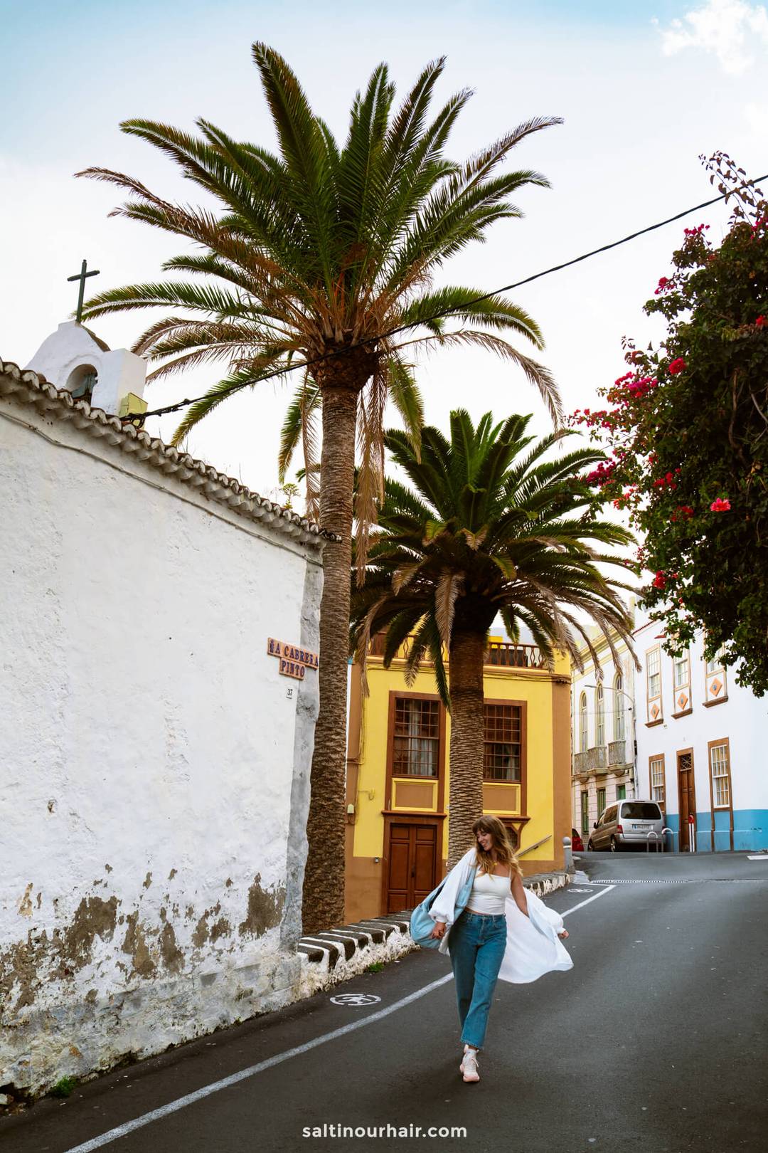 A woman in jeans and a white shirt walks on a narrow street lined with palm trees and colorful buildings in La Palma, Canary Islands, with a church visible on the left&mdash;a perfect scene for any Travel Guide.