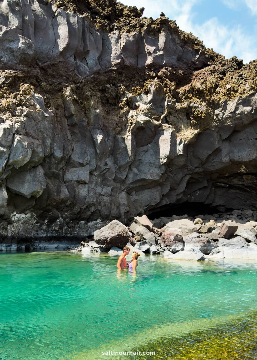 Two people stand in clear turquoise water near rocky cliffs and a small cave under a partly cloudy sky, capturing the natural beauty of La Palma in the Canary Islands&mdash;a perfect scene for any travel guide.
