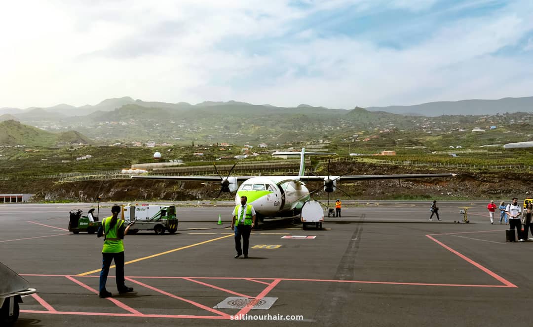 A small passenger plane is parked on an airport tarmac in La Palma, with ground crew and equipment nearby; green hills and houses are visible in the background under a partly cloudy Canary Islands sky.