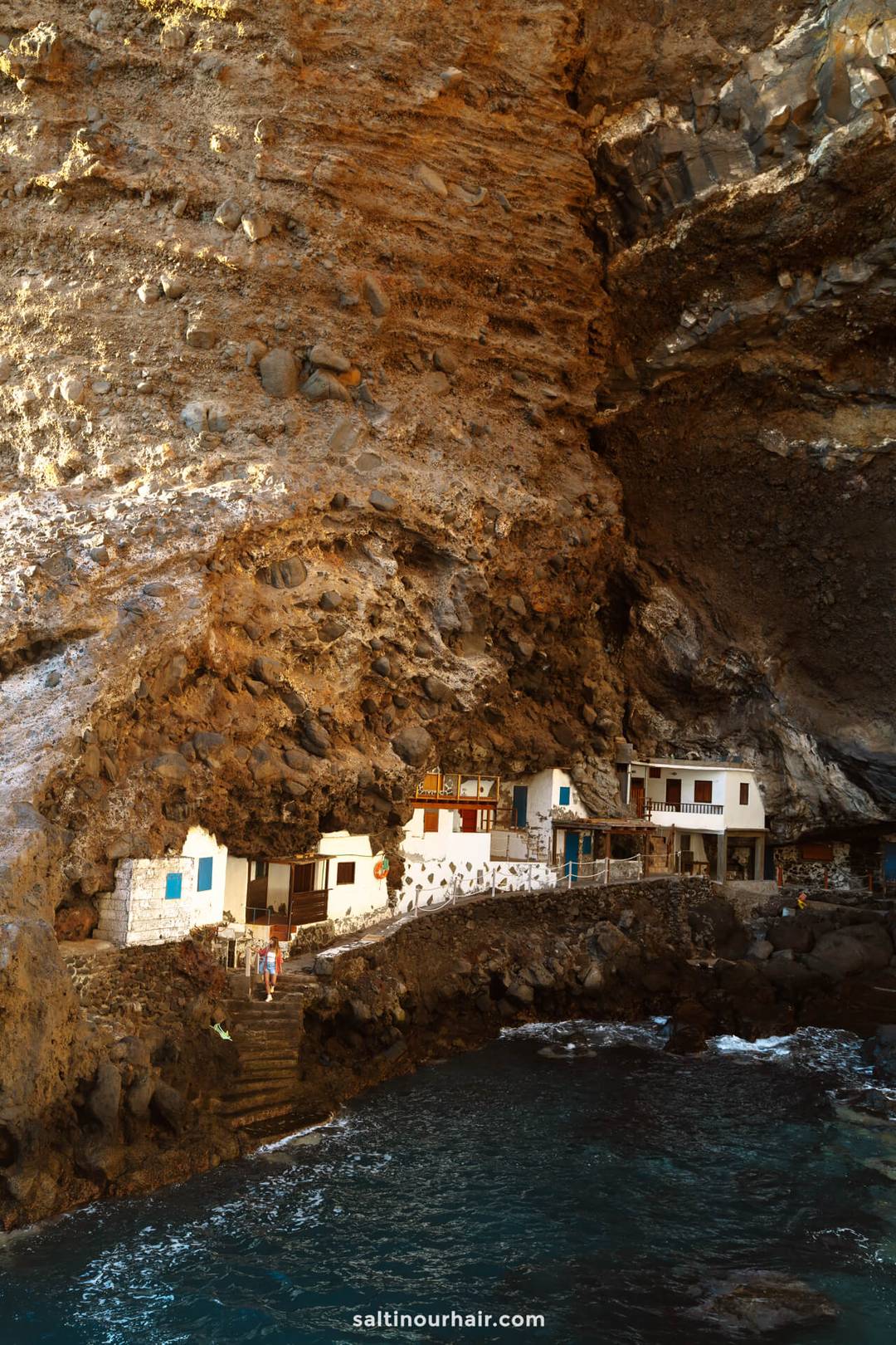 Small white houses built into a large rocky cave overlook blue ocean water on La Palma in the Canary Islands, with stairs leading down to the shore&mdash;a hidden gem for any travel guide.