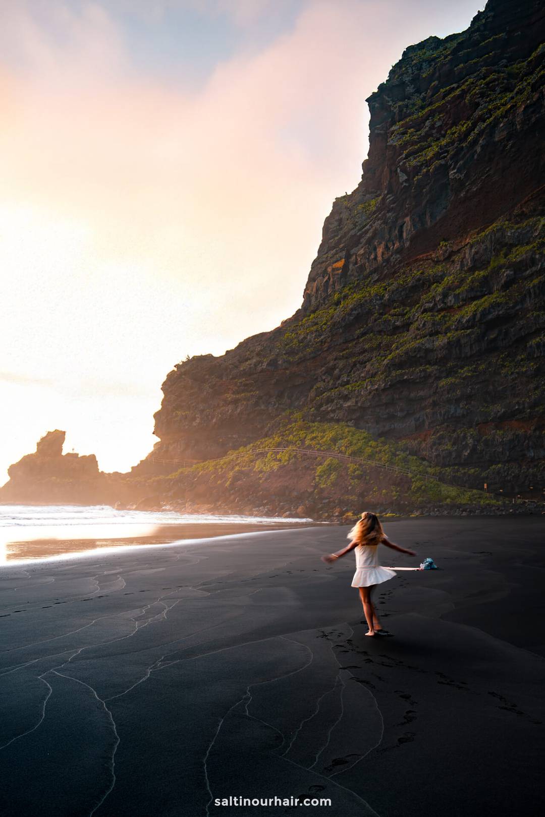 A woman in a white dress stands on a black sand beach near tall cliffs at sunset in La Palma, Canary Islands, her footprints trailing behind&mdash;an inspiring scene for any travel guide.