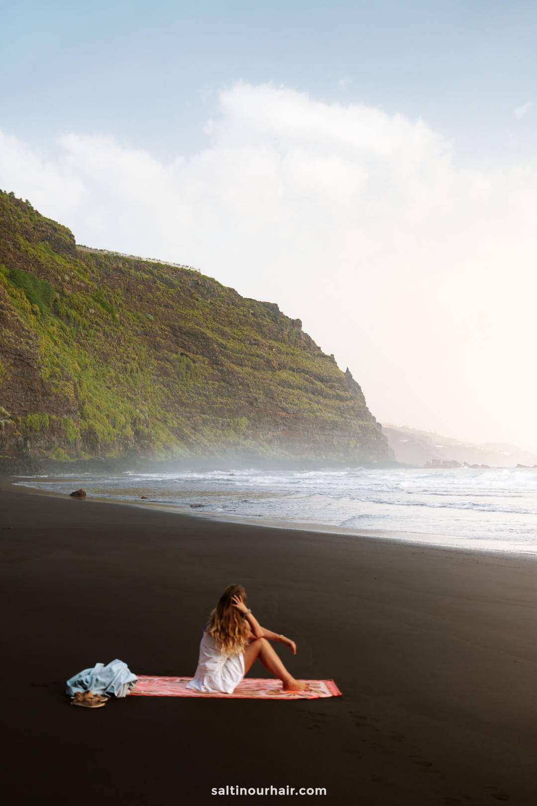 A woman sits on a towel on a black sand beach in La Palma near the ocean, with green cliffs in the background under a clear sky&mdash;a perfect moment from any Canary Islands travel guide.