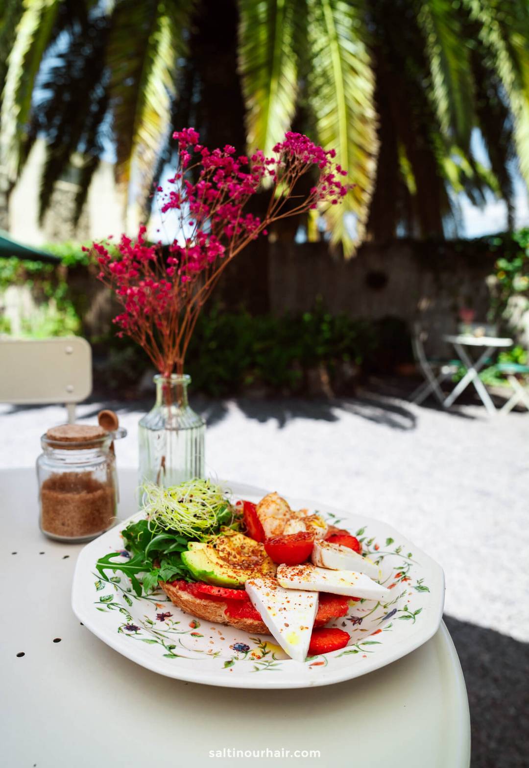 A plate of sliced cheese, tomatoes, avocado, and greens on toast sits on a white table outdoors in La Palma, with a jar of brown sugar and a vase of pink flowers beside it&mdash;perfect for a Canary Islands travel guide.