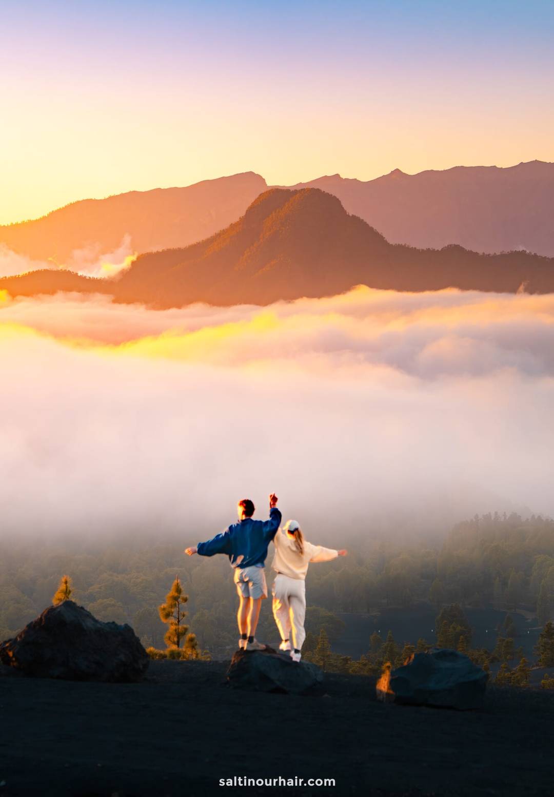 Two people stand on rocks overlooking a foggy valley in La Palma, Canary Islands, with mountains in the background during sunrise, wearing casual clothes and facing the view&mdash;an inspiring moment from a Travel Guide.