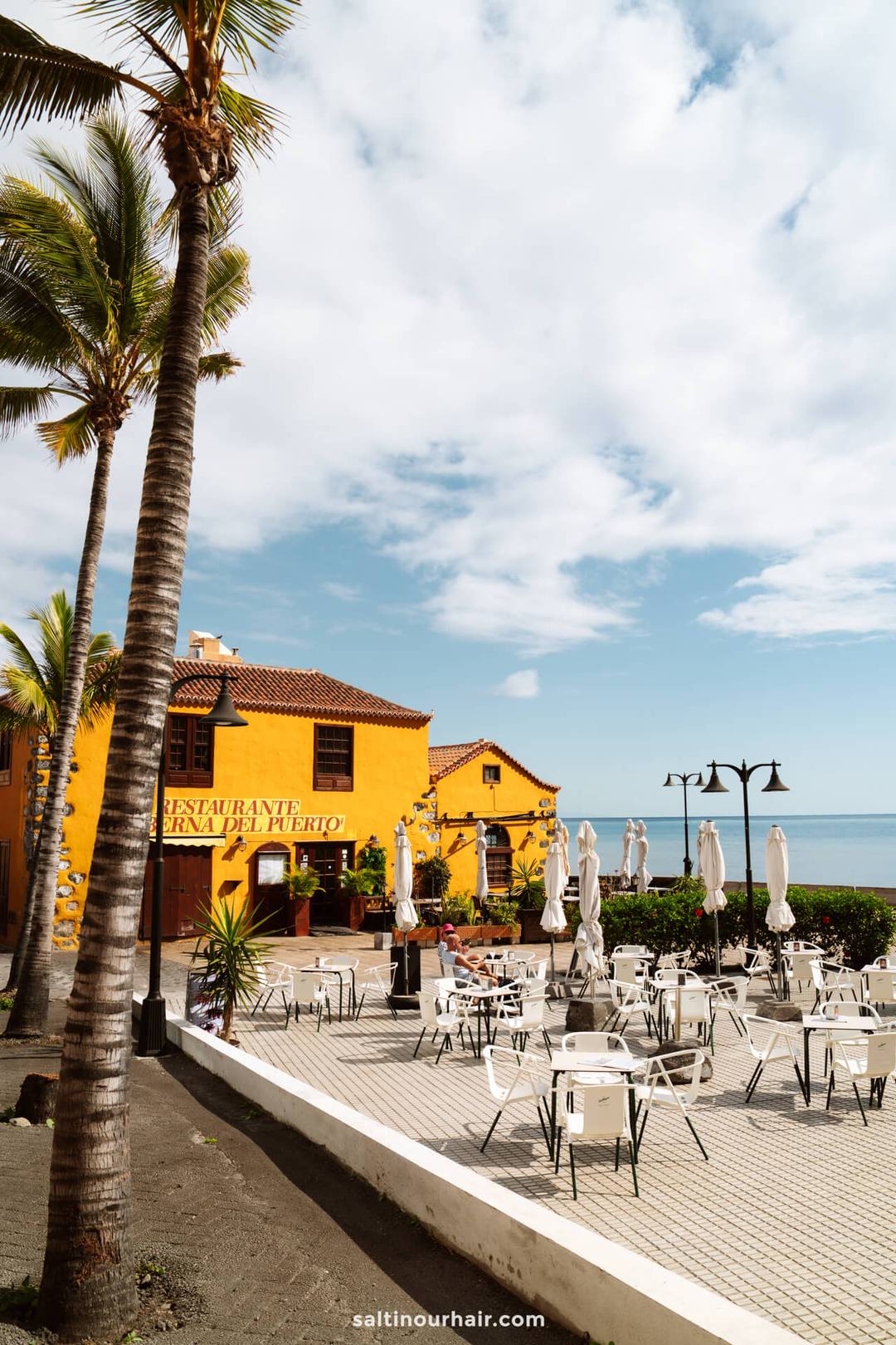 Outdoor restaurant patio with empty white tables and chairs, palm trees, and a yellow building near the ocean under a partly cloudy sky&mdash;an inviting La Palma scene from a Canary Islands travel guide.