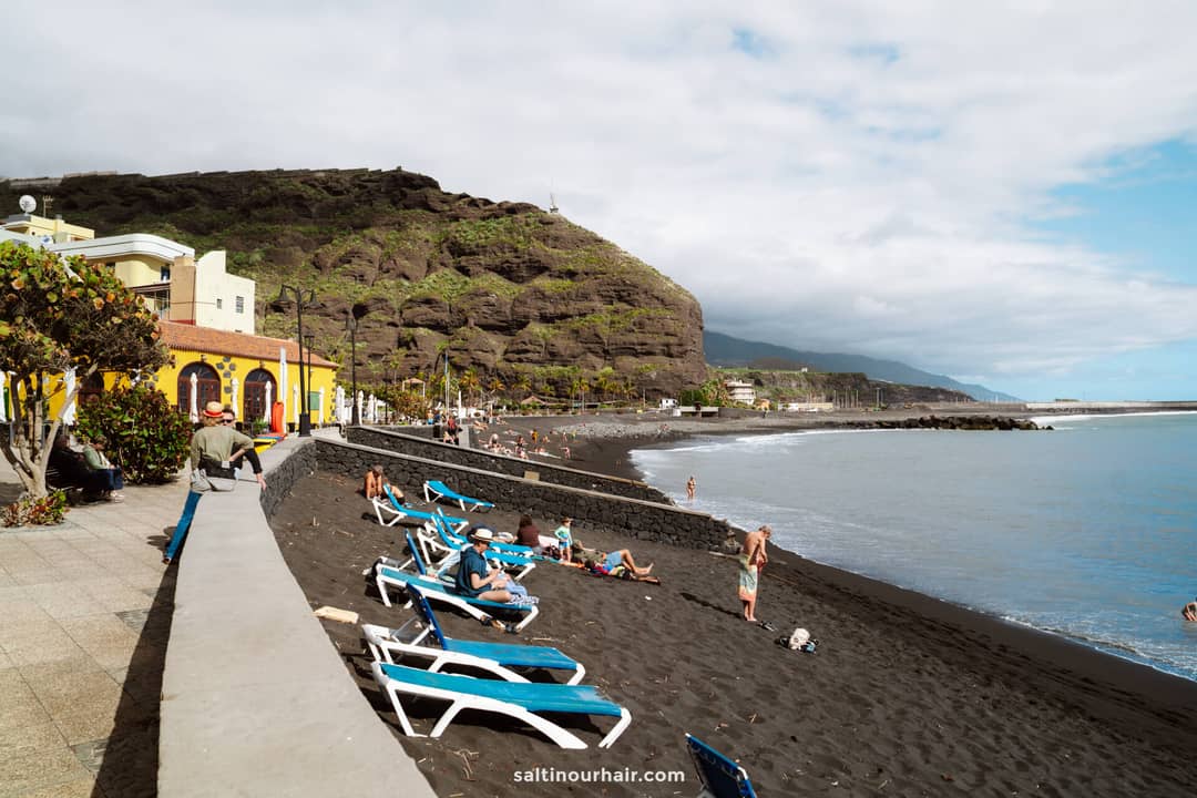 People relax on blue lounge chairs on a black sand beach in La Palma, bordered by a stone wall, with cliffs and colorful buildings in the background under a partly cloudy sky&mdash;a perfect scene from the Canary Islands Travel Guide.
