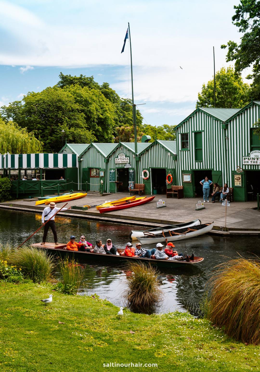 A group of people rides in a punt boat on a canal near green-and-white striped boat sheds, with kayaks and people on the dock&mdash;a classic scene to include on your New Zealand South Island itinerary.