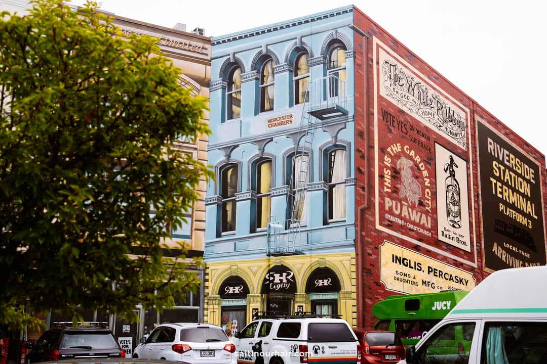 A lively street scene reminiscent of stops on a New Zealand South Island itinerary, with colorful historic buildings adorned with painted advertisements, parked cars, and a leafy tree in the foreground.