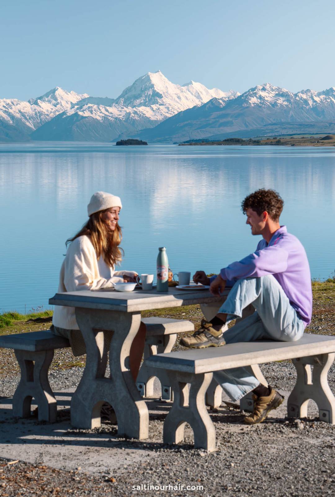Two people sit at a concrete picnic table by a lake, enjoying a meal beneath snow-capped mountains and clear blue sky&mdash;a perfect pause on any New Zealand South Island itinerary.