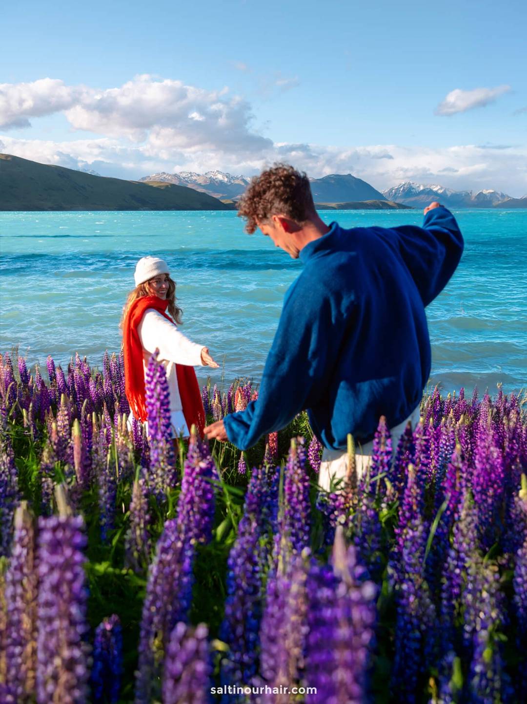 Two people stand among purple lupines by a turquoise lake, with mountains and a partly cloudy sky in the background&mdash;a scene straight out of a New Zealand South Island itinerary. One person faces the camera, reaching toward the other.