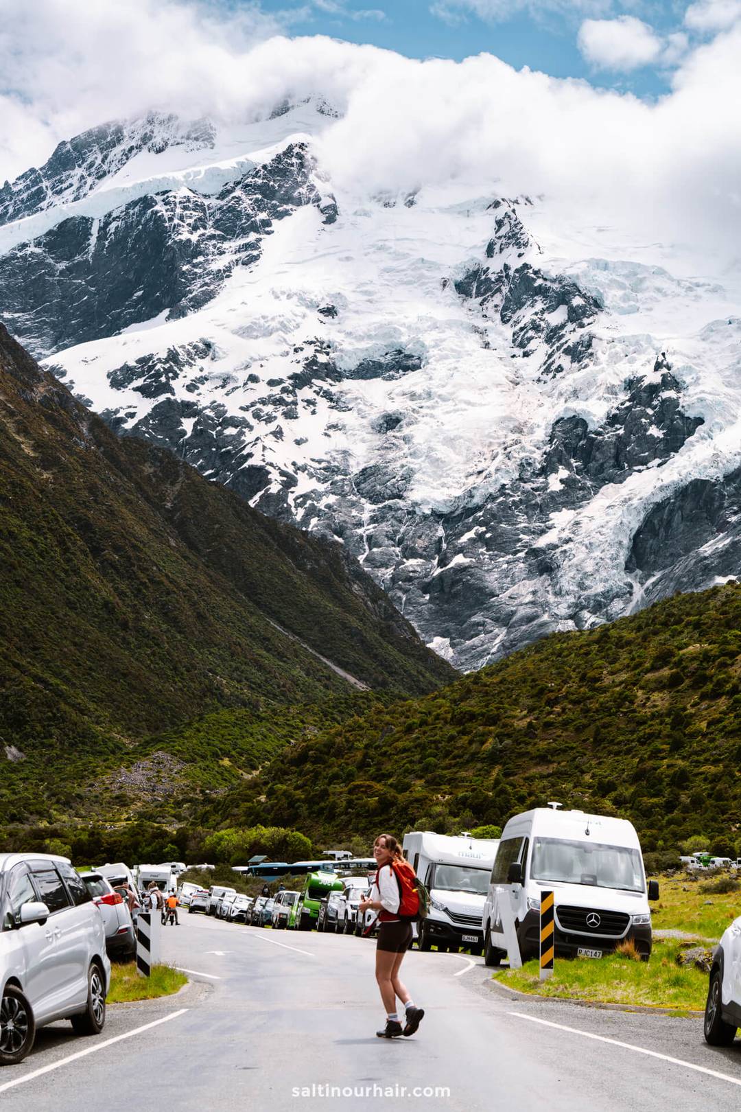 A woman with a backpack walks along a mountain road lined with parked cars and vans, framed by snow-covered peaks and clouds&mdash;a perfect scene for any New Zealand South Island itinerary.