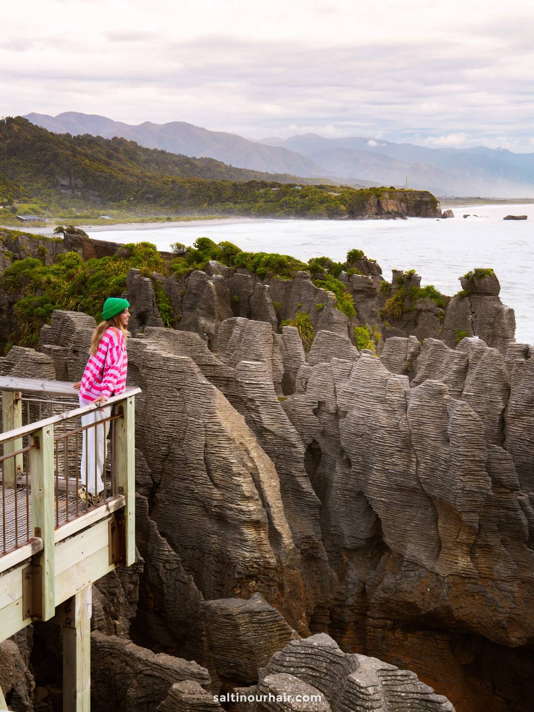 A person in a pink and white striped top stands on a viewing platform overlooking the Pancake Rocks at Punakaiki&mdash;an essential stop on any New Zealand South Island itinerary&mdash;with the ocean and mountains in the background.