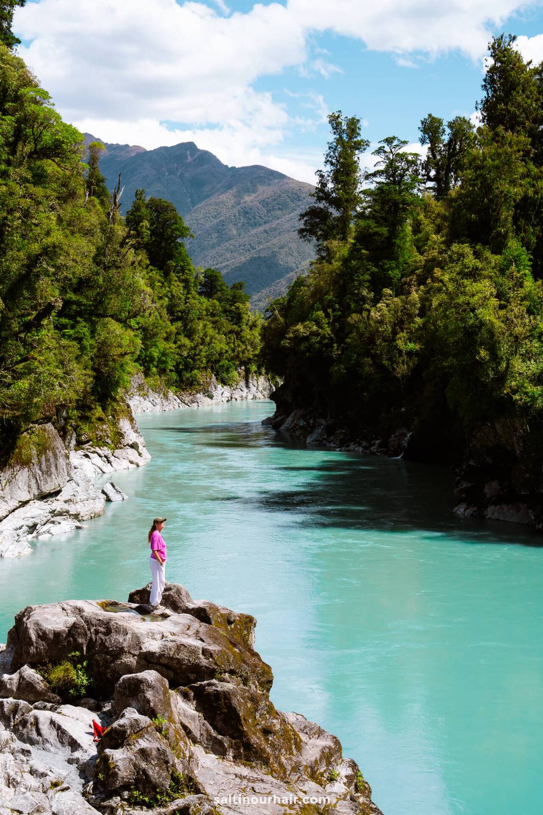 A person in a pink shirt stands on a rocky outcrop above a turquoise river winding through a forested valley with mountains in the background, capturing the essence of any New Zealand South Island itinerary.
