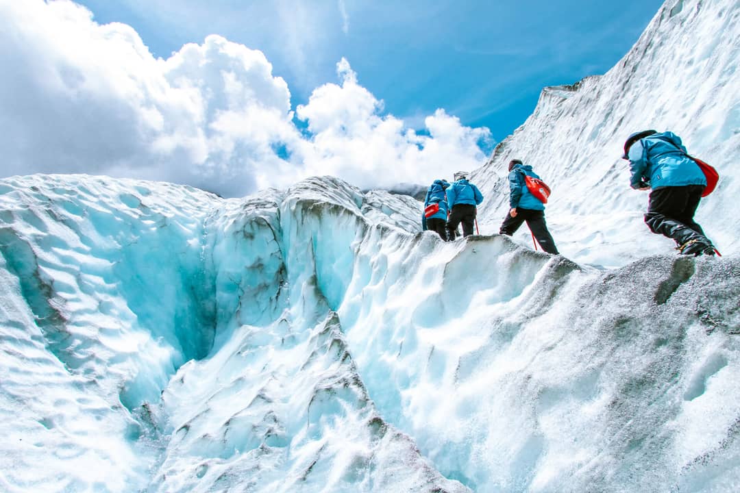 Three hikers in blue jackets and red backpacks trek across a glacier