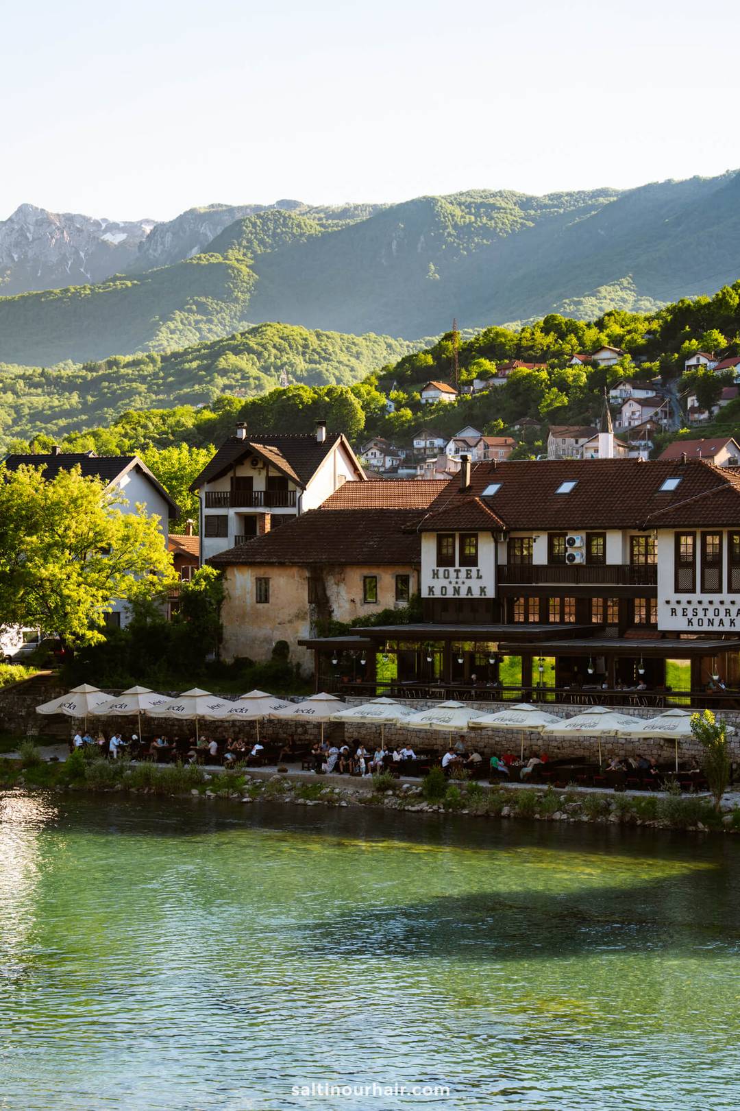 Riverside hotel with outdoor seating, surrounded by green hills and mountains, with several houses in the background&mdash;an ideal stop on your Bosnia itinerary for a sunny day.
