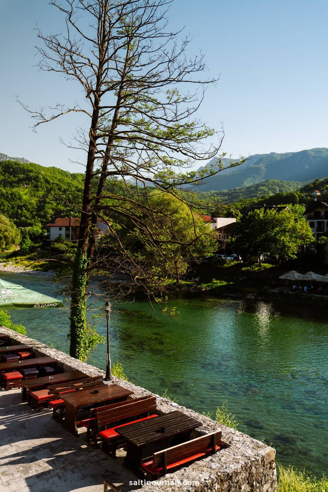 Outdoor seating with wooden benches faces a calm, clear river&mdash;an inviting stop on any Bosnia itinerary. A leafless tree stands nearby, framed by green hills and houses under a clear blue sky.
