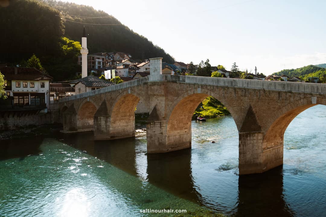 Stone arch bridge crosses a clear river with a village and mosque in the background, surrounded by green hills under sunlight&mdash;a must-see highlight on any Bosnia itinerary.