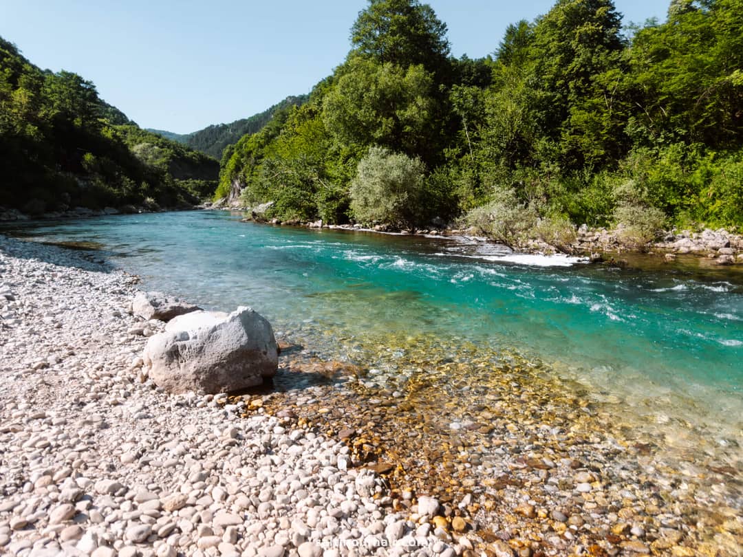 Clear turquoise river flows through a rocky, pebbled shore surrounded by dense green trees under a clear blue sky&mdash;a must-see spot on any Bosnia itinerary.