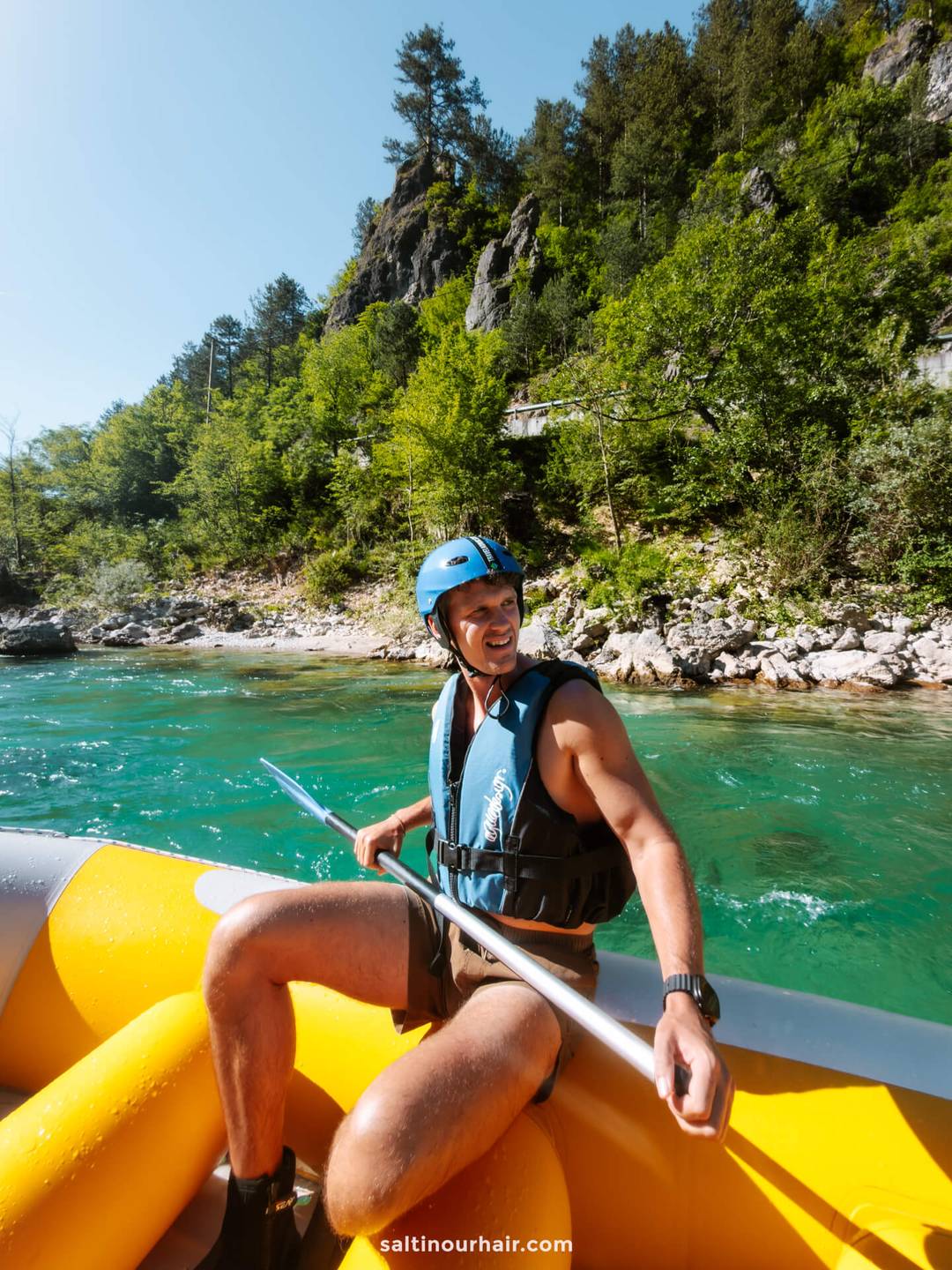 A man wearing a helmet and life vest sits in a yellow inflatable raft on a river, holding a paddle, surrounded by trees and rocky terrain&mdash;perfect for adding adventure to your Bosnia itinerary.