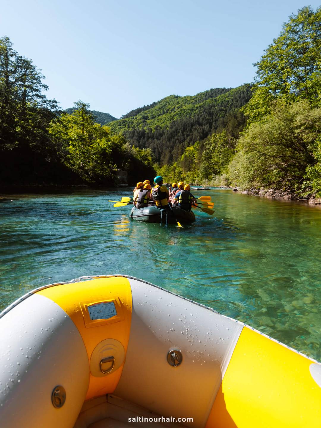 Two inflatable rafts with people wearing helmets and life jackets paddle down a clear, green river surrounded by lush trees and hills under a blue sky&mdash;a perfect adventure on any Bosnia itinerary.