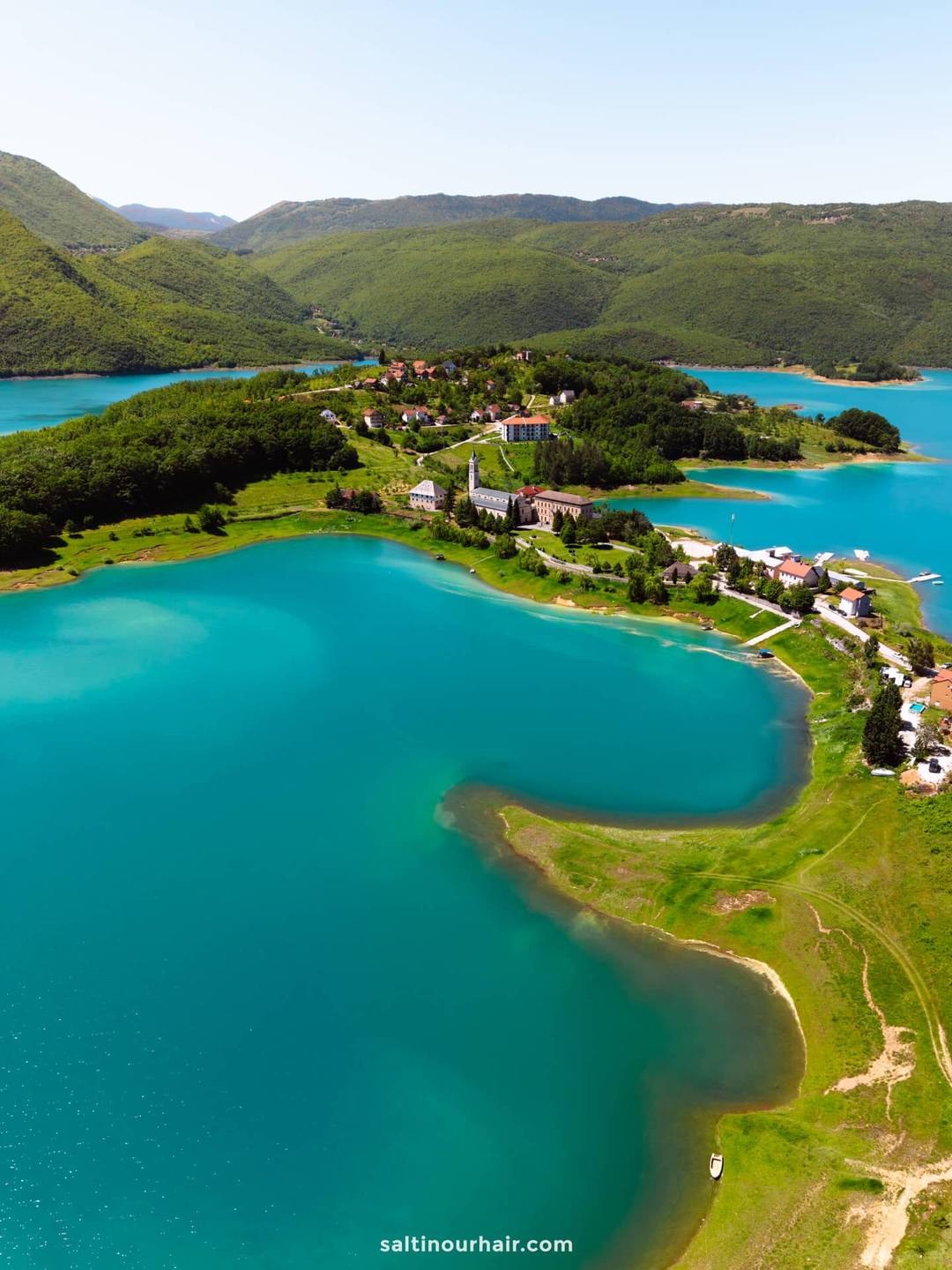 Aerial view of a turquoise lake surrounded by green hills and a small village with scattered buildings near the shoreline, a perfect highlight for any Bosnia itinerary.