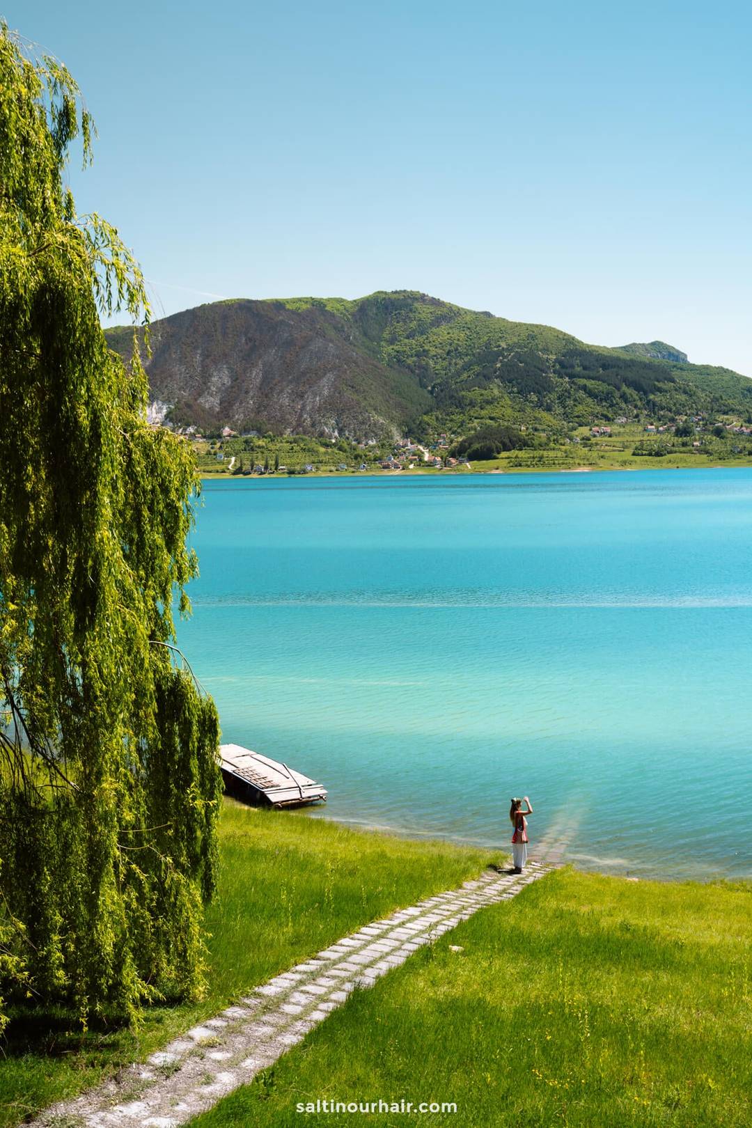 A person stands at the end of a stone path by a turquoise lake&mdash;a peaceful stop that belongs on any Bosnia itinerary&mdash;with a small dock, green grass, and mountains in the background under a clear blue sky.